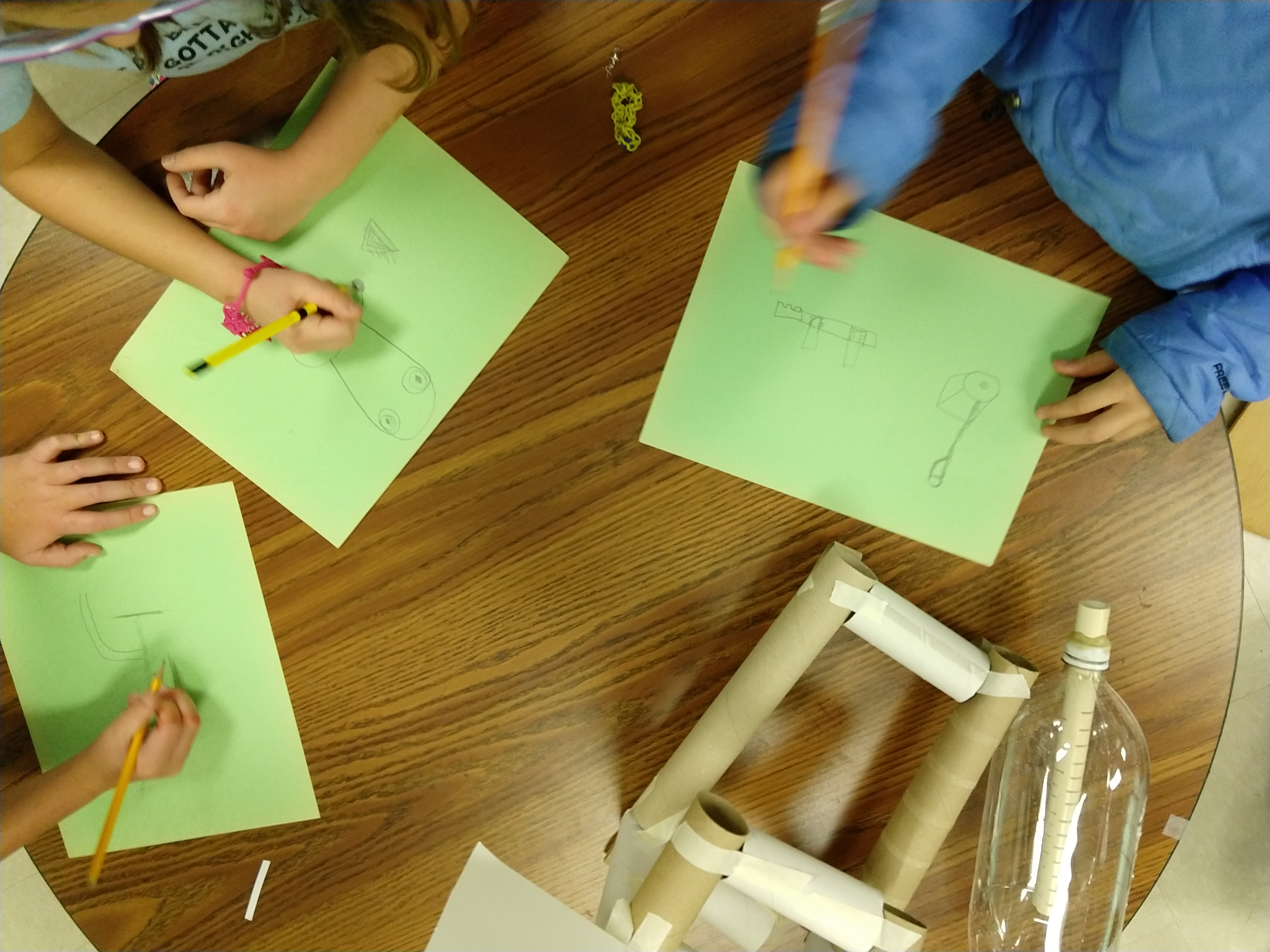 Photo of students working on activities at a table, including sketching design ideas on paper and building creations using recyclables and masking tape.